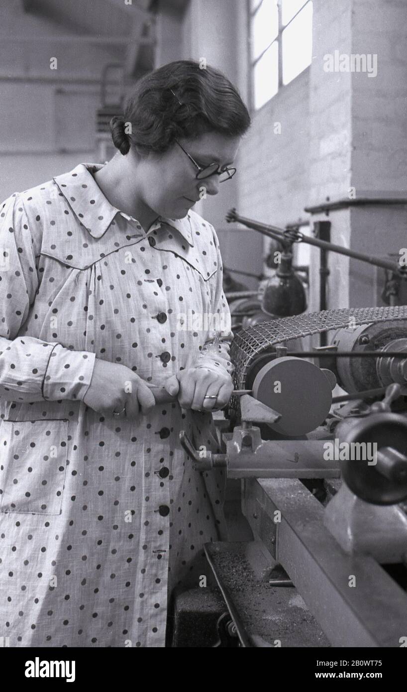 Anni '1940, storica, una signora in una classe di scuola notturna di lavorazione del legno, che indossa un tabard floreale a maniche lunghe o un soprabito, a un banco di lavoro utilizzando una macchina utensile per macinare o affilare uno strumento metallico utilizzato nell'artigianato, in Inghilterra, nel Regno Unito. Foto Stock