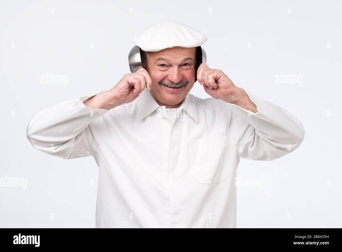 Chef ispanico senior che copre le orecchie con padelle da cucina su sfondo bianco Foto Stock
