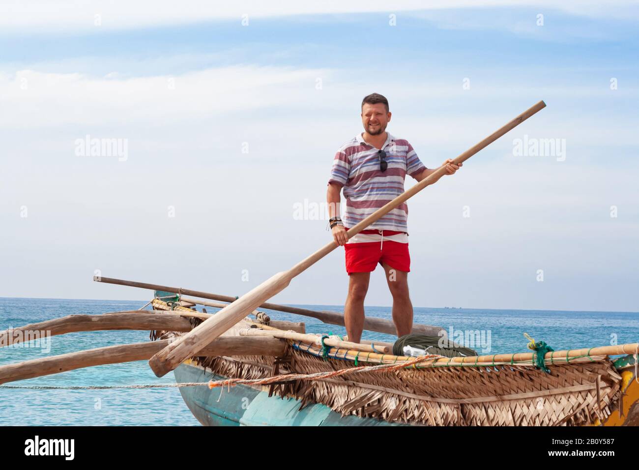 Ritratto di un uomo con una barba media in una camicia e pantaloncini rossi su una tradizionale barca di pescatori con una pagaia nelle sue mani. Foto Stock