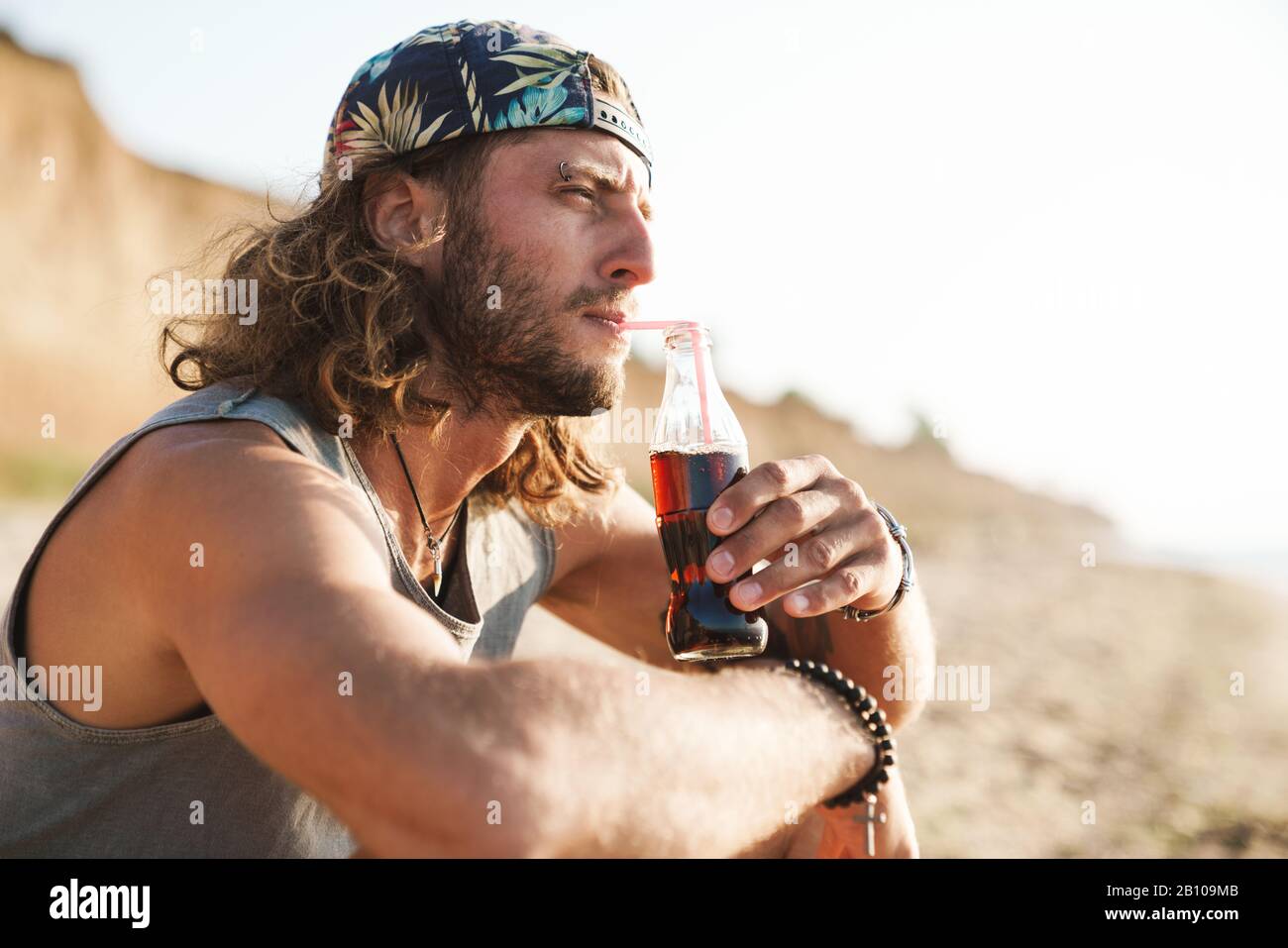 Foto di pensare giovane uomo indossare con capelli ricci bere soda e seduta sulla spiaggia soleggiata Foto Stock