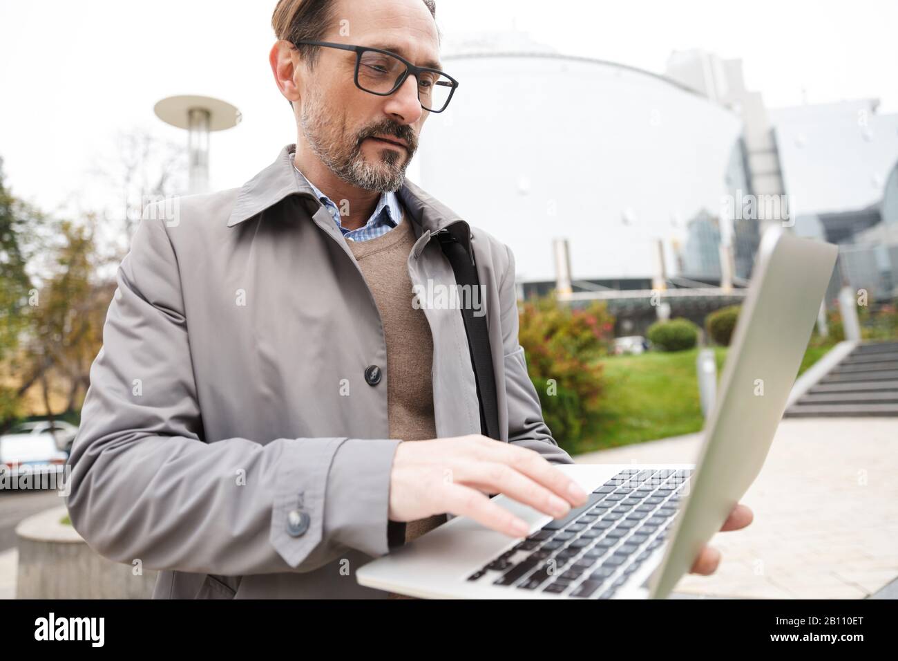 Immagine di un uomo d'affari concentrato con bearded in occhiali usando un laptop mentre si sta in piedi in strada della città Foto Stock