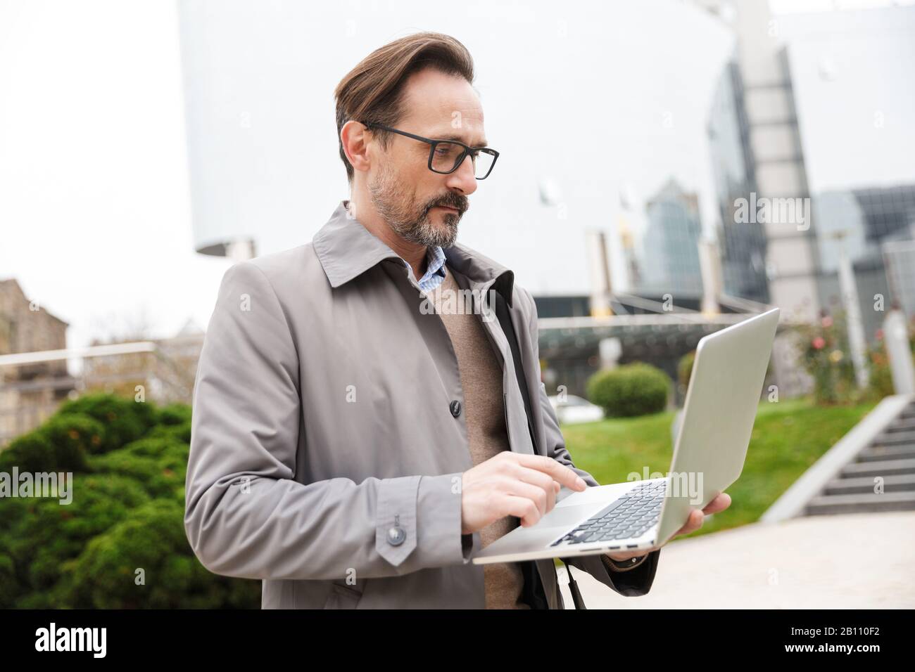 Immagine di un uomo d'affari concentrato in occhiali usando un computer portatile mentre si trova in città strada Foto Stock
