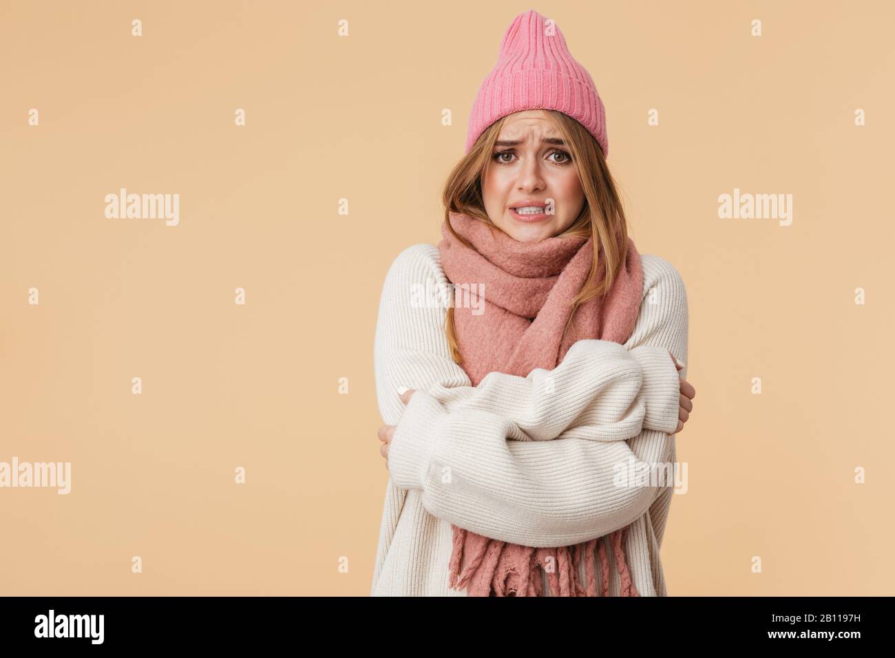 Immagine della giovane ragazza caucasica che indossa cappello invernale e sciarpa tremante e sensazione di freddo isolato su sfondo beige Foto Stock