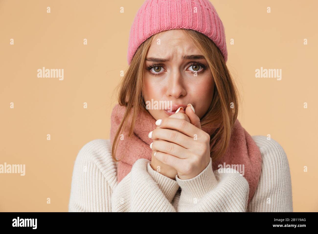 Immagine della giovane ragazza caucasica che indossa cappello invernale e sciarpa tremante e sensazione di freddo isolato su sfondo beige Foto Stock