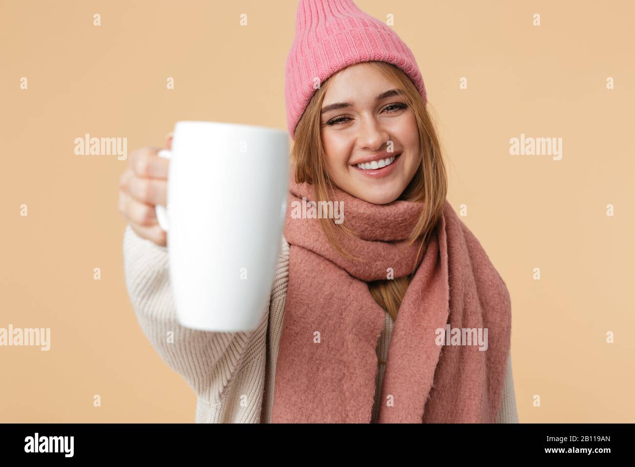 Immagine di giovane ragazza attraente in cappello invernale sorridente e tenendo la tazza isolata su sfondo beige Foto Stock