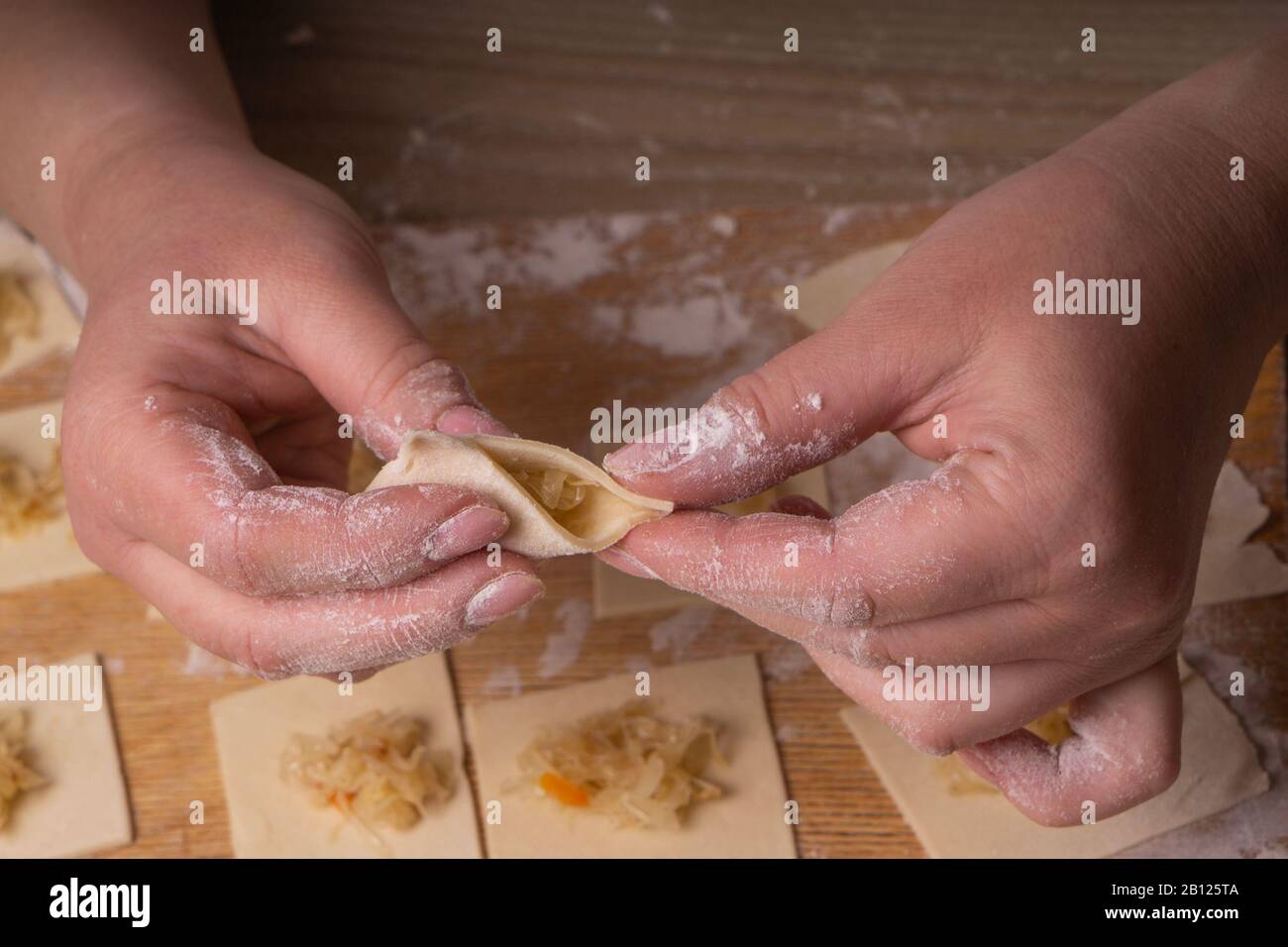 Una donna scolpisce gnocchi e ravioli da quadrati di pasta e cavolo. Pannello di taglio compensato, setaccio di farina di legno e perno di laminazione di legno - attrezzi per Foto Stock