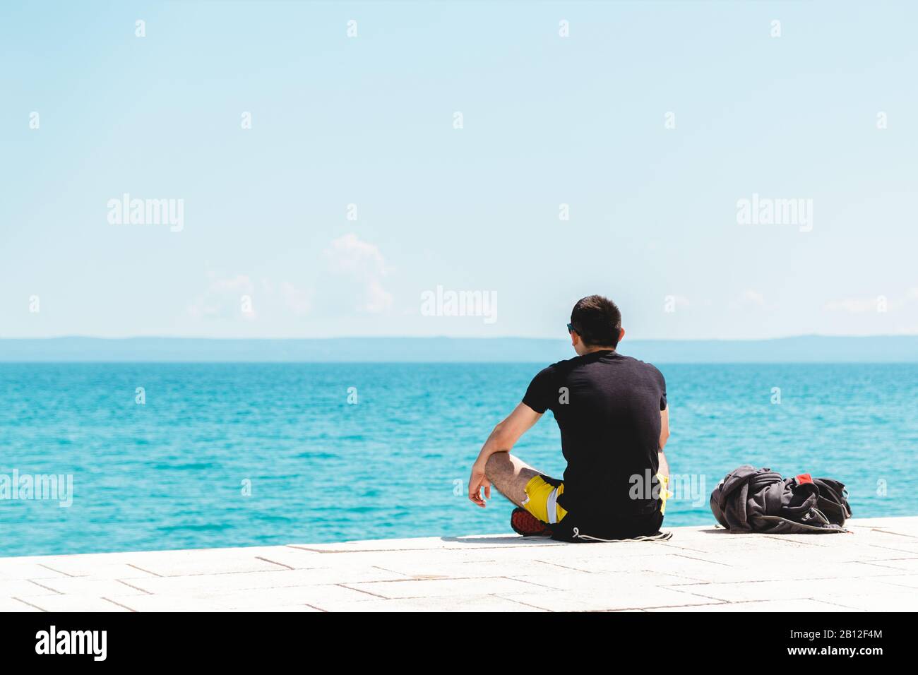 giovane uomo viaggiatore solo con zaino vicino a lui meditando guardando l'oceano e godendo il silenzio Foto Stock