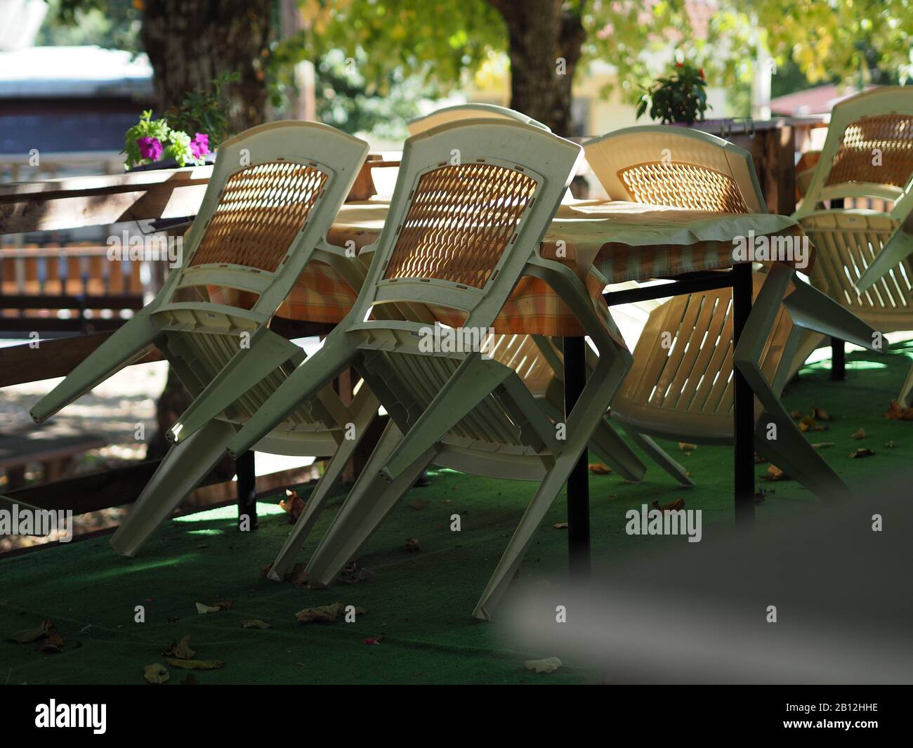 Sedie appoggiate contro un tavolo e gettate ombre in una taverna in Macedonia, Grecia, con un albero coperto piazza dietro. Foto Stock