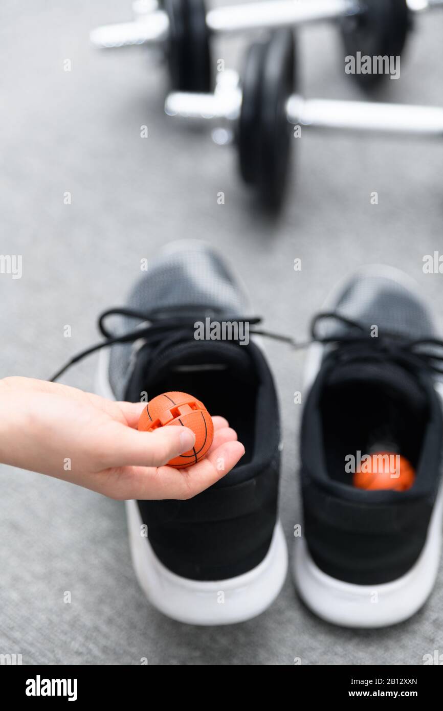 Mano che tiene una palla di deodorante di scarpa su scarpe da allenamento con manubri sullo sfondo. Concetto per evitare l'odore sgradevole nelle calzature Foto Stock