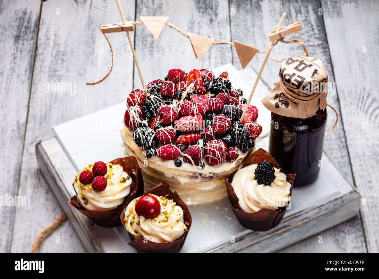 Torta di frittelle con un berretto di frutti di bosco inzuppato in cioccolato bianco su un tavolo bianco di legno con alcuni muffin belgi e una bottiglia di sciroppo Foto Stock
