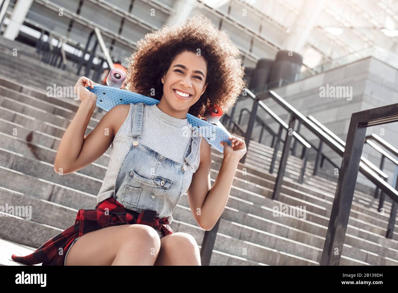 Giovane donna nella strada della città seduta su scale con Penny board guardando macchina fotografica confusa Foto Stock