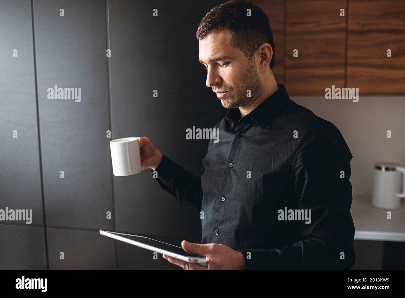 Vista laterale del giovane uomo d'affari in cucina, lavora in remoto. Imprenditore tenere tablet e guardare. Bere il tè durante la pausa caffè. Lavoro efficiente a casa. Foto Stock