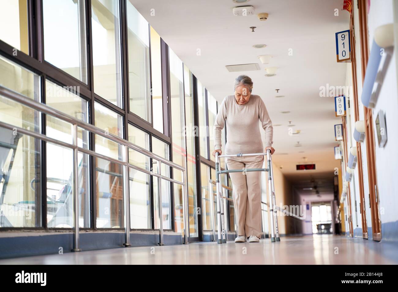 donna anziana asiatica che cammina usando un camminatore nel senso del corridoio di casa di cura, vista ad angolo basso. Foto Stock