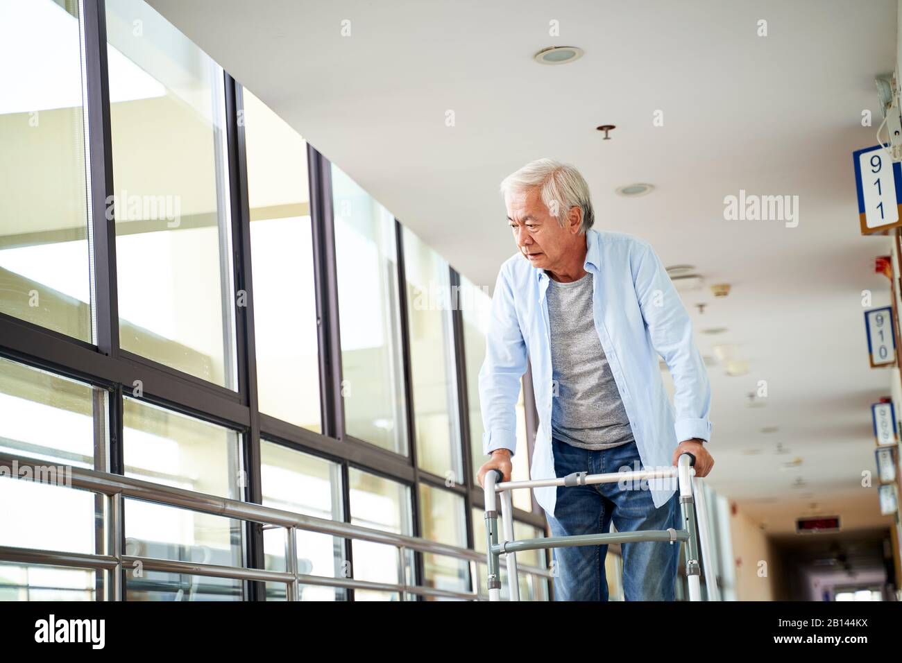 l'uomo anziano aisiano che cammina con difficoltà usando un camminatore nel corridoio della sede di cura Foto Stock