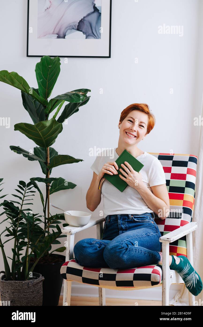 Donna con i capelli corti dello zenzero seduta in una sedia a casa con un libro nelle mani Foto Stock