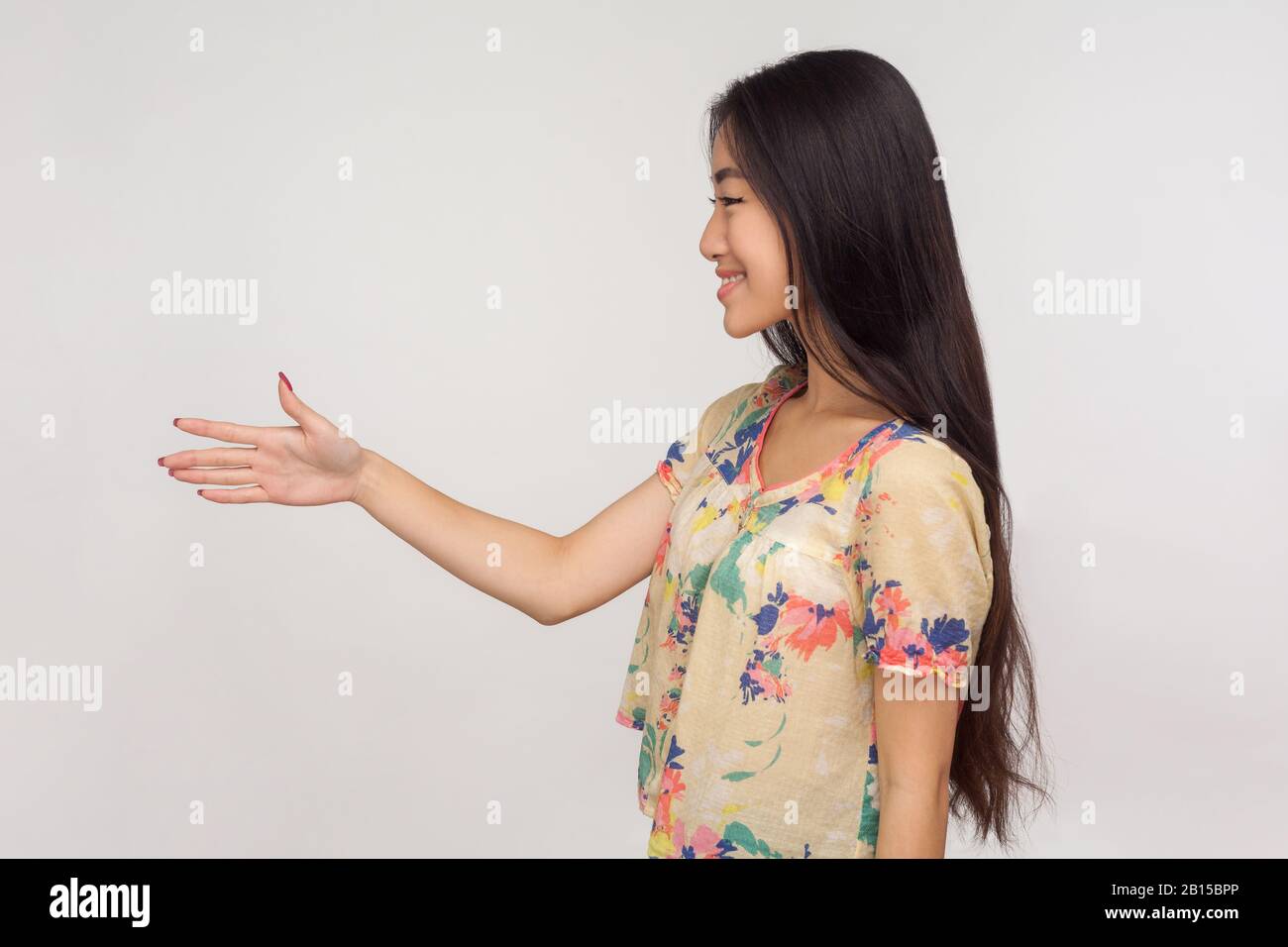 Vista laterale della ragazza asiatica amichevole con lunghi capelli brunette in blusa dando mano a stretta di mano e sorridente al cliente, saluto e familiarizzarsi. In Foto Stock