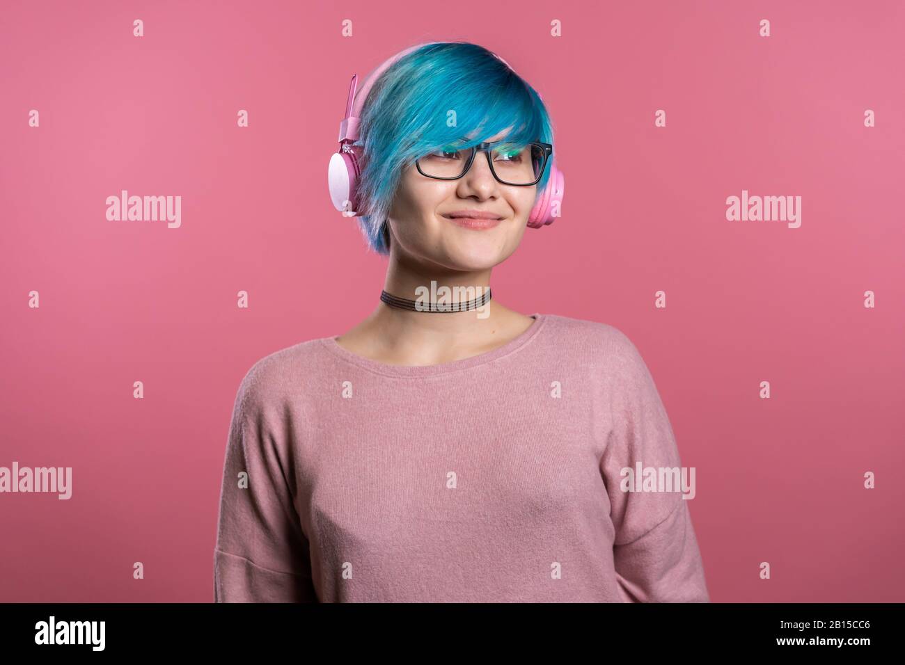Bella ragazza con capelli blu che si divertono, sorridono, ballando con cuffie rosa in studio su sfondo colorato. Musica, danza, concetto radio. Foto Stock
