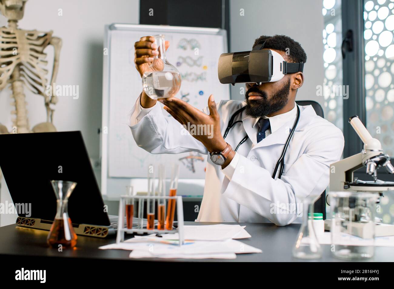 Giovane medico africano, scienziato, virologo, indossando occhiali per la realtà virtuale per l'immaginazione, conducendo ricerche in laboratorio, tenendo matraccio in mano Foto Stock