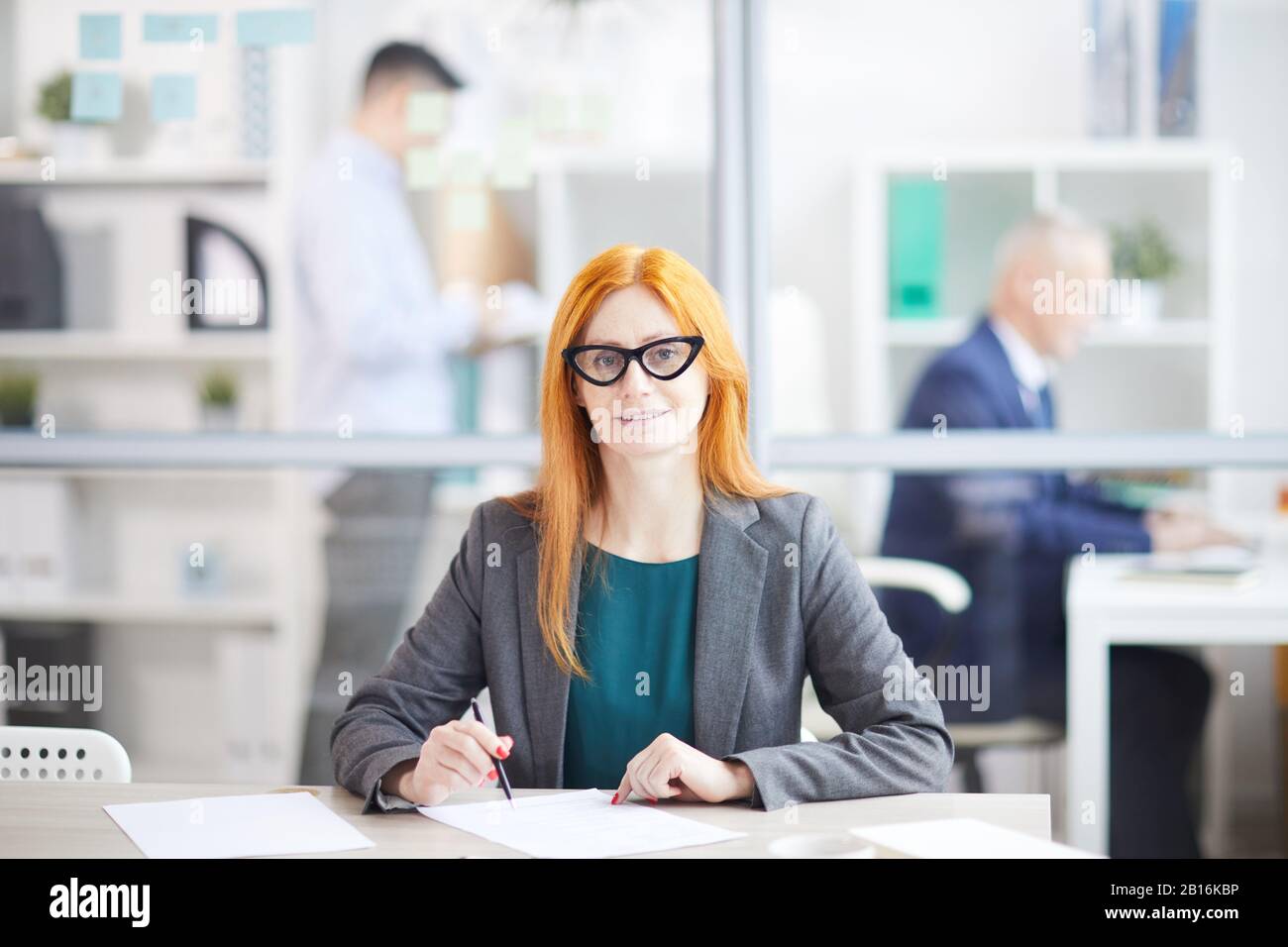 Ritratto di donna d'affari rossa adulta sorridente alla macchina fotografica mentre posa sul posto di lavoro in ufficio cubicolo, copia spazio Foto Stock