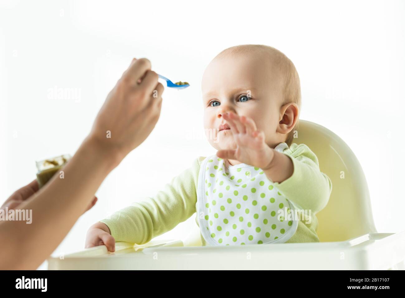Fuoco selettivo del bambino cute che tira la mano alla madre con il cucchiaio di nutrizione del bambino isolato sul bianco Foto Stock