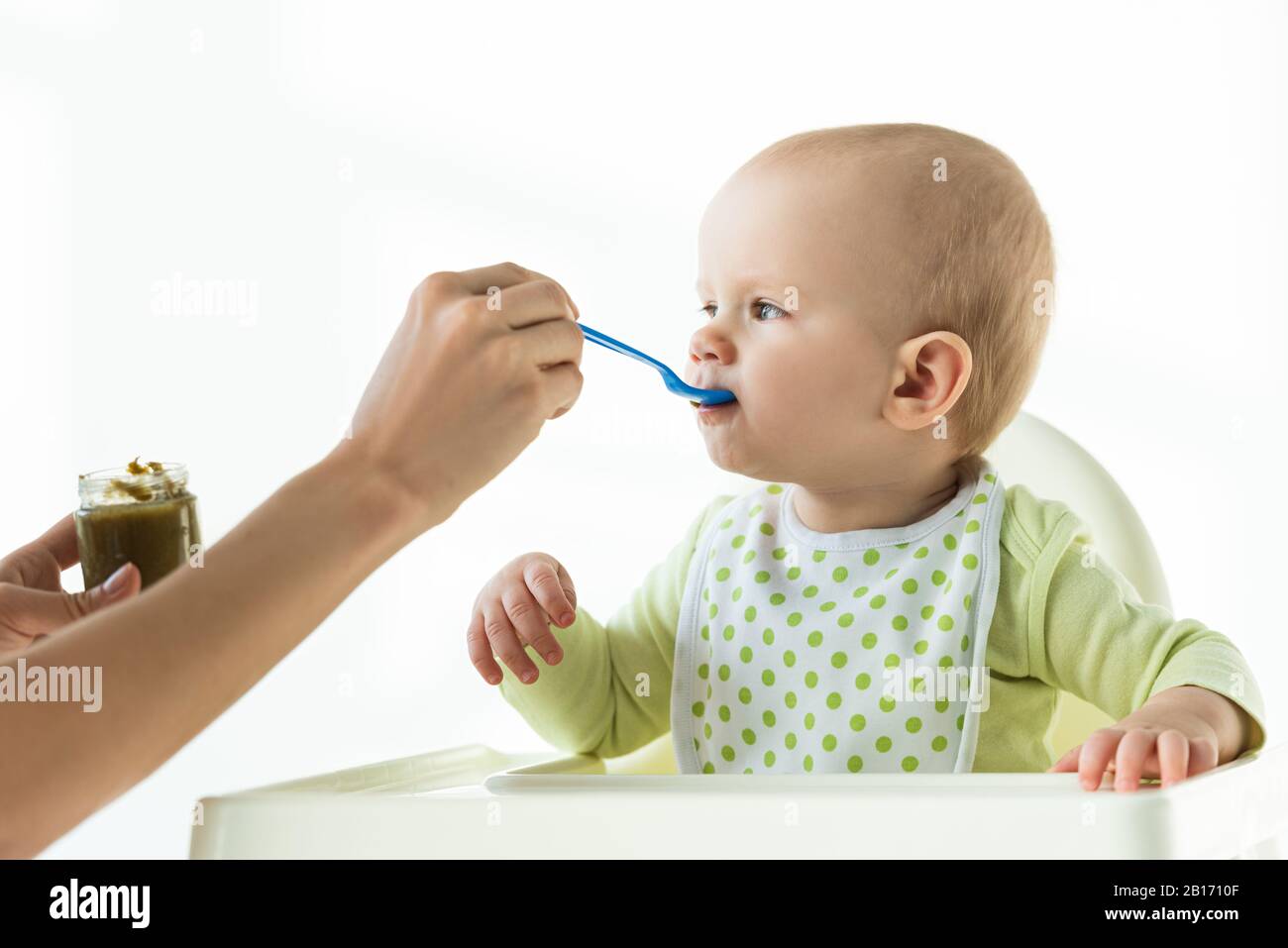 Madre con vasetto di nutrizione del bambino e cucchiaio che alimenta il bambino su sedia di alimentazione su sfondo bianco Foto Stock