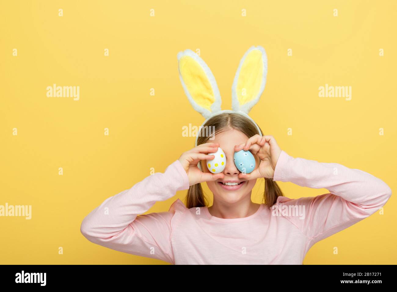 bambino felice con orecchie conigliate che coprono gli occhi con uova di pasqua isolate su giallo Foto Stock