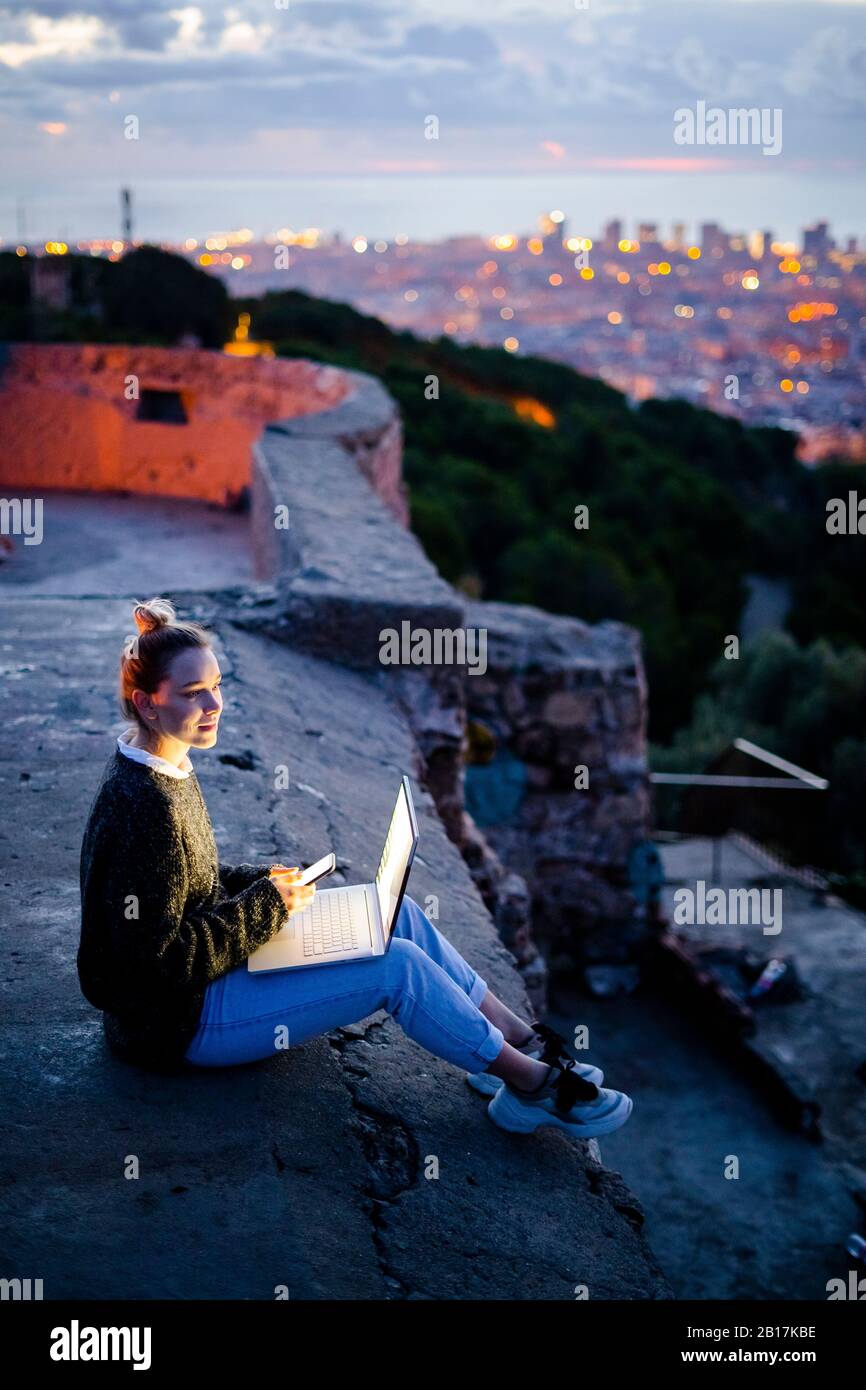 Giovane donna seduta sulla ringhiera di protezione al di sopra della città utilizzando il telefono cellulare, Barcellona, Spagna Foto Stock