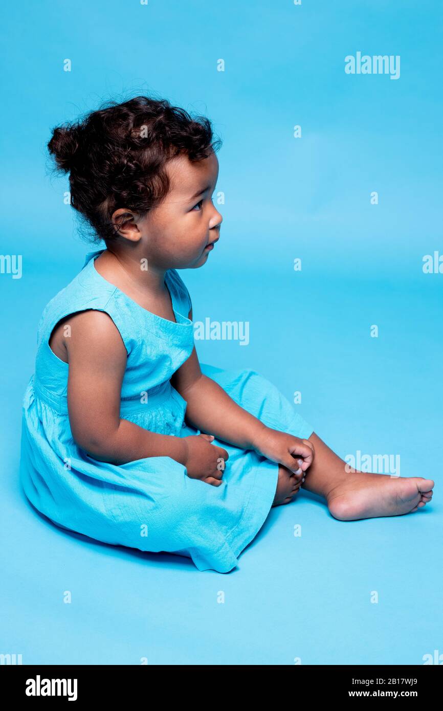 Bambina a piedi nudi che indossa un abito azzurro seduta su sfondo blu Foto Stock