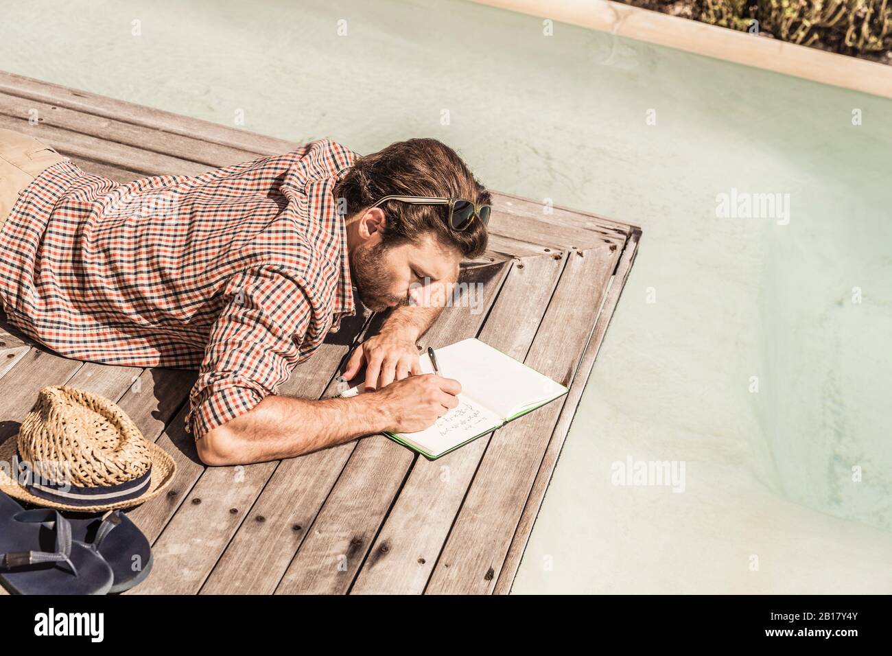 Uomo che giace accanto a una piscina che scrive in un notebook Foto Stock