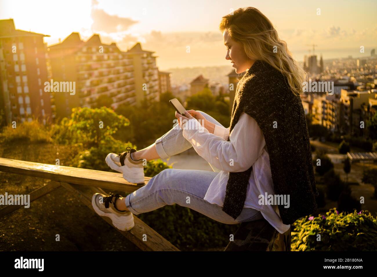 Giovane donna seduta sulla ringhiera di protezione al di sopra della città utilizzando il telefono cellulare, Barcellona, Spagna Foto Stock