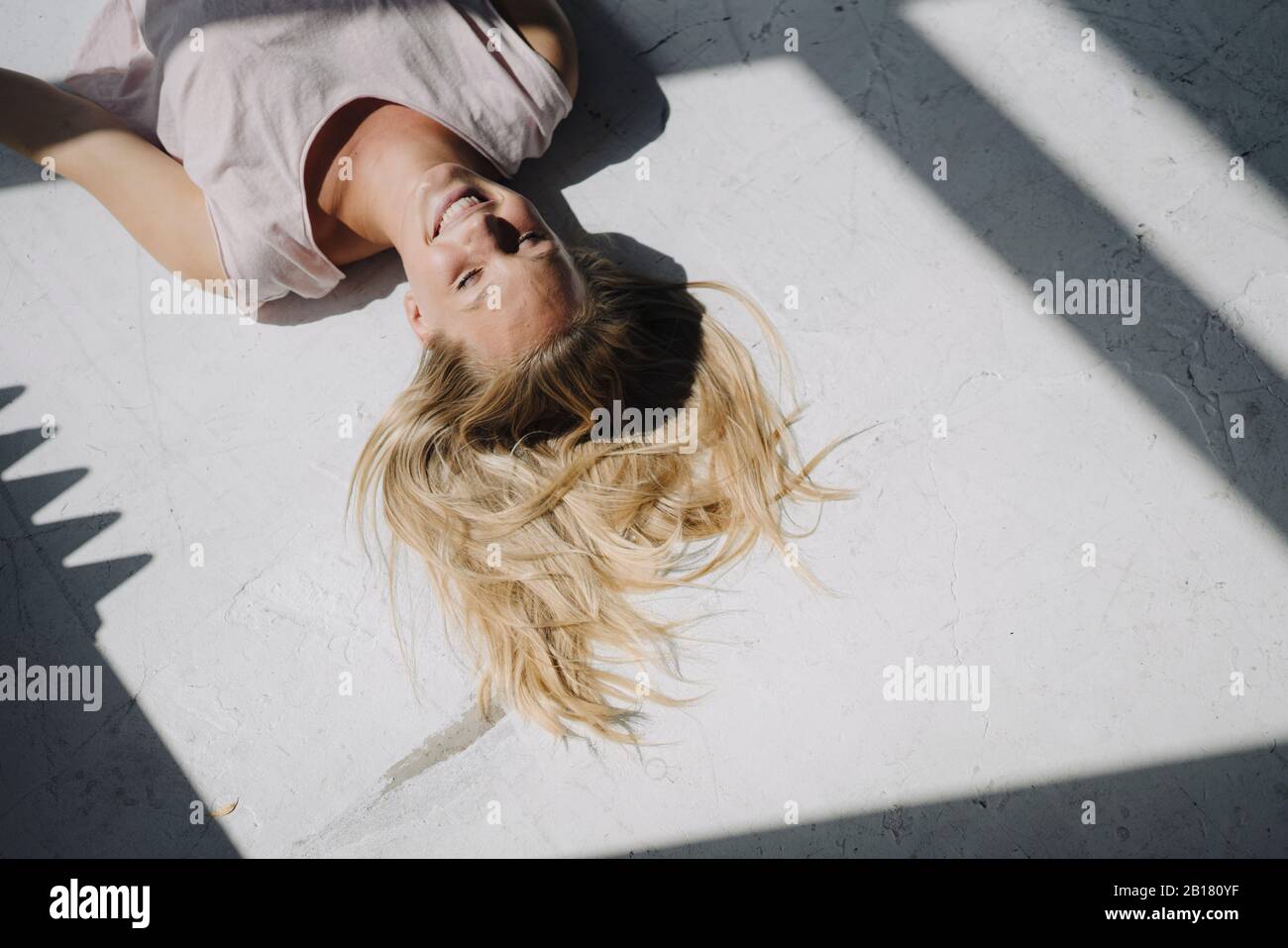 Giovane donna bionda sorridente sdraiata sul pavimento alla luce del sole Foto Stock