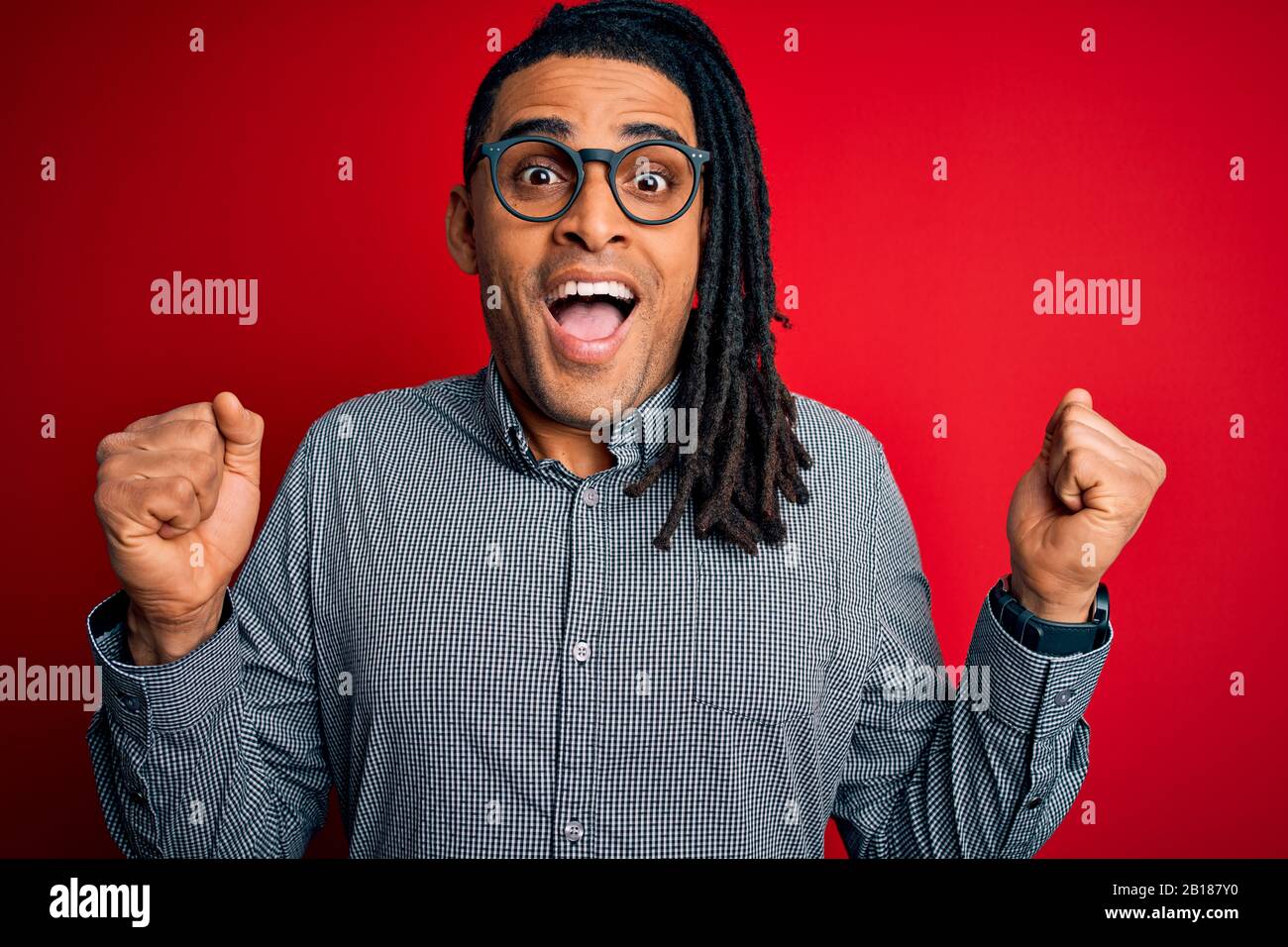 Giovane bell'uomo afroamericano con temute serrature che indossano camicia casual e occhiali che celebrano sorpreso e stupito per il successo con le braccia sollevate e. Foto Stock
