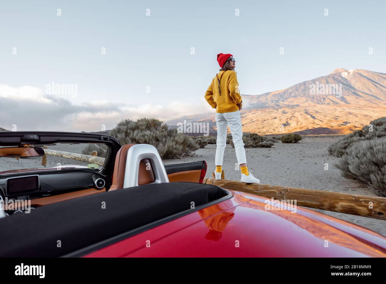 Donna vestita di maglione luminoso e cappello che gode di grandi paesaggi di montagna mentre si sta in piedi sulla recinzione stradale. Viaggiando in auto sull'isola di Tenerife, vista panoramica Foto Stock