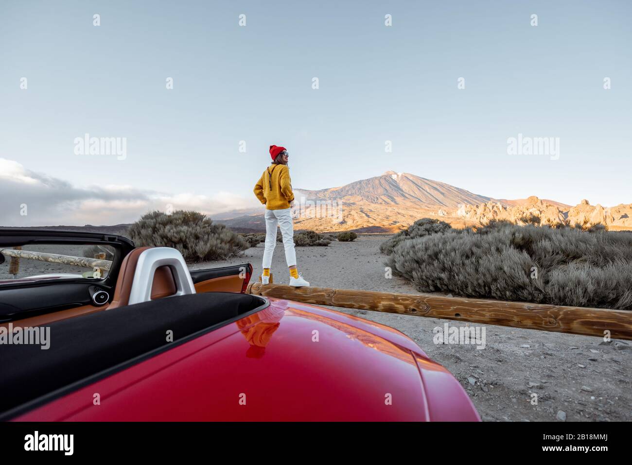 Donna vestita di maglione luminoso e cappello che gode di grandi paesaggi di montagna mentre si sta in piedi sulla recinzione stradale. Viaggiando in auto sull'isola di Tenerife, vista panoramica Foto Stock