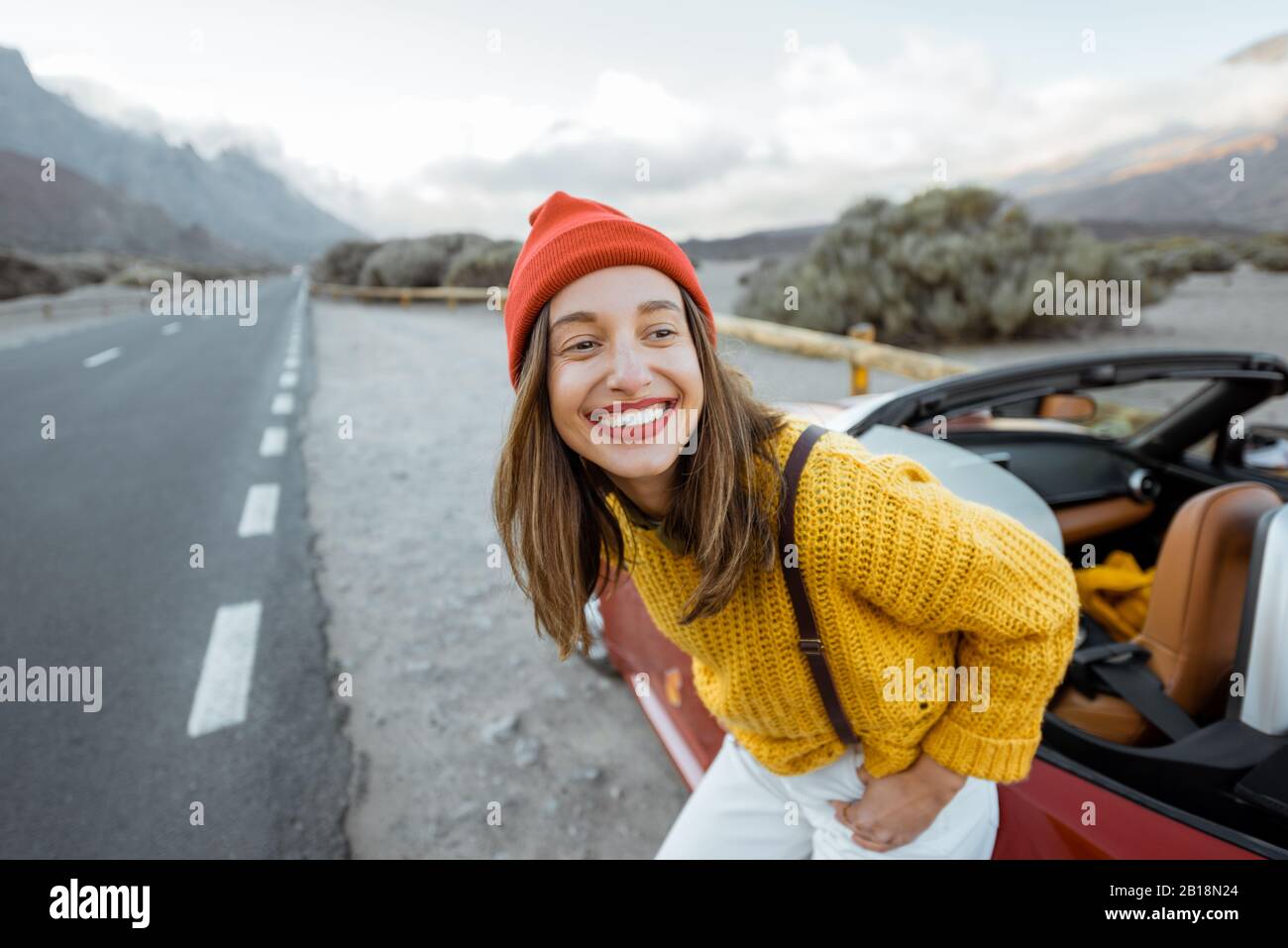 Ritratto di una donna gioiosa vestita di maglione luminoso e cappello che si gode viaggio su strada, in piedi vicino alla macchina sulla strada della pittoresca valle vulcanica su un tramonto Foto Stock