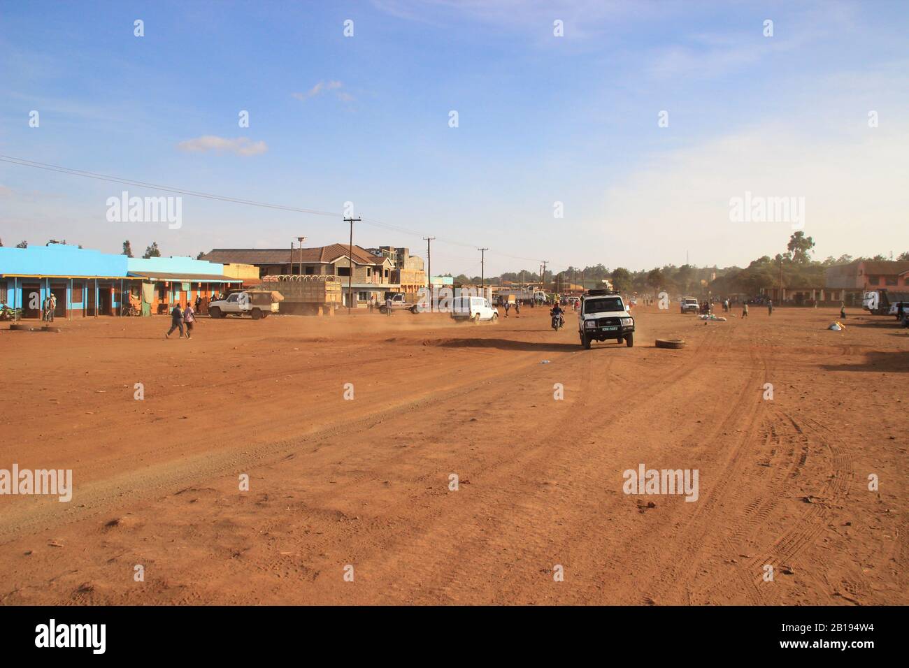 Marsabit, Kenya - 16 gennaio 2015: Una strada cittadina con una strada coperta d'arancia, negozi e un'auto Foto Stock