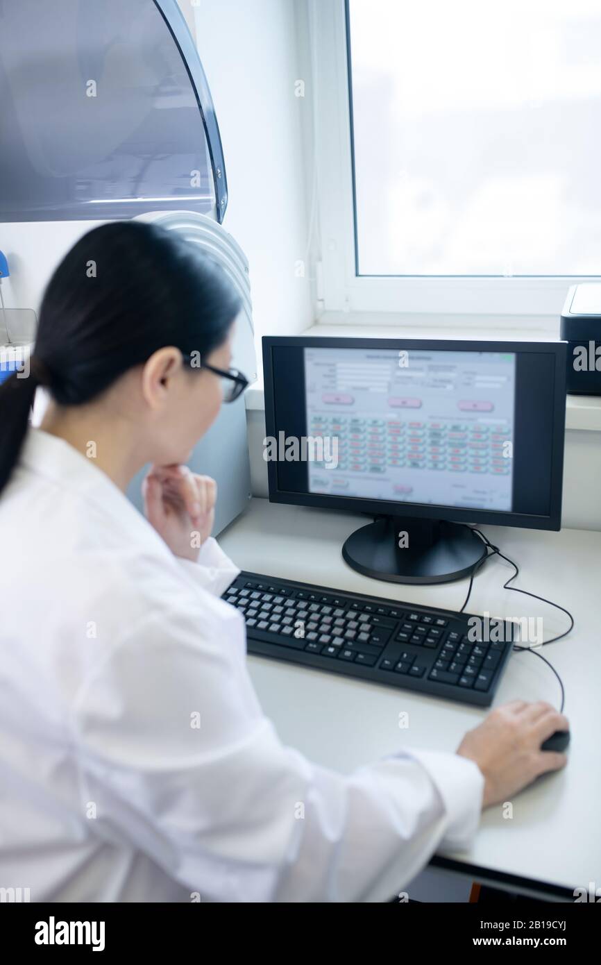 Dottore dai capelli scuri in un abito bianco che lavora su un computer che guarda pensieroso Foto Stock