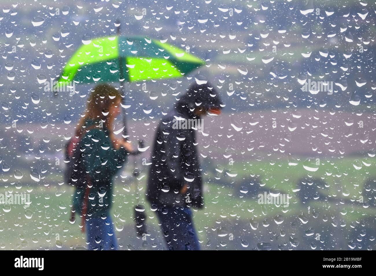 Riassunto di una coppia che cammina sotto la pioggia con un ombrello, visto attraverso una finestra coperta di goccioline di pioggia. Wet tempo UK. Foto Stock
