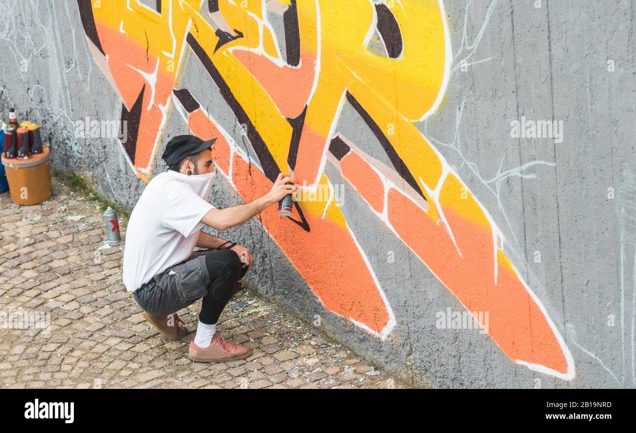 Graffiti artista che copre il suo viso durante la pittura con spray a colori sul muro - Urbano, Street art, generazione millennial, murale concetto - Focus on hi Foto Stock