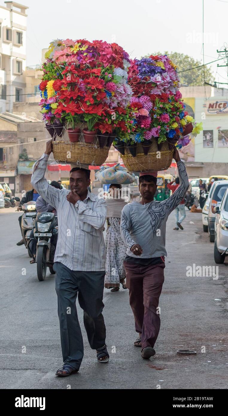 Jamnagar, Gujarat/India - Dicembre 18 2018: Due uomini camminano su una strada in cemento trafficata che porta grandi cestini di fiori colorati sulla loro testa. Foto Stock