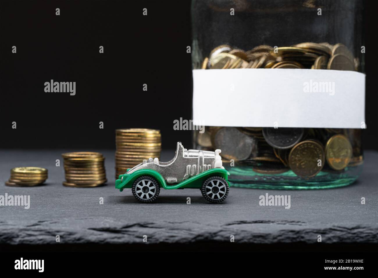 Pile di monete, vasetto con etichetta bianca e un'auto giocattolo. Salvataggio per un concetto astratto del veicolo. Copiare lo spazio a sinistra. Foto Stock