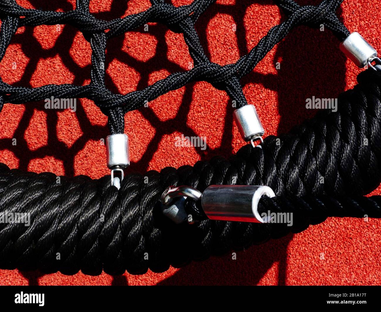 dettaglio swing in vinile nero di forma rotonda in un parco giochi. pavimento di sicurezza in gomma morbida granulare rossa sottostante. forti ombre astratte alla luce del sole Foto Stock