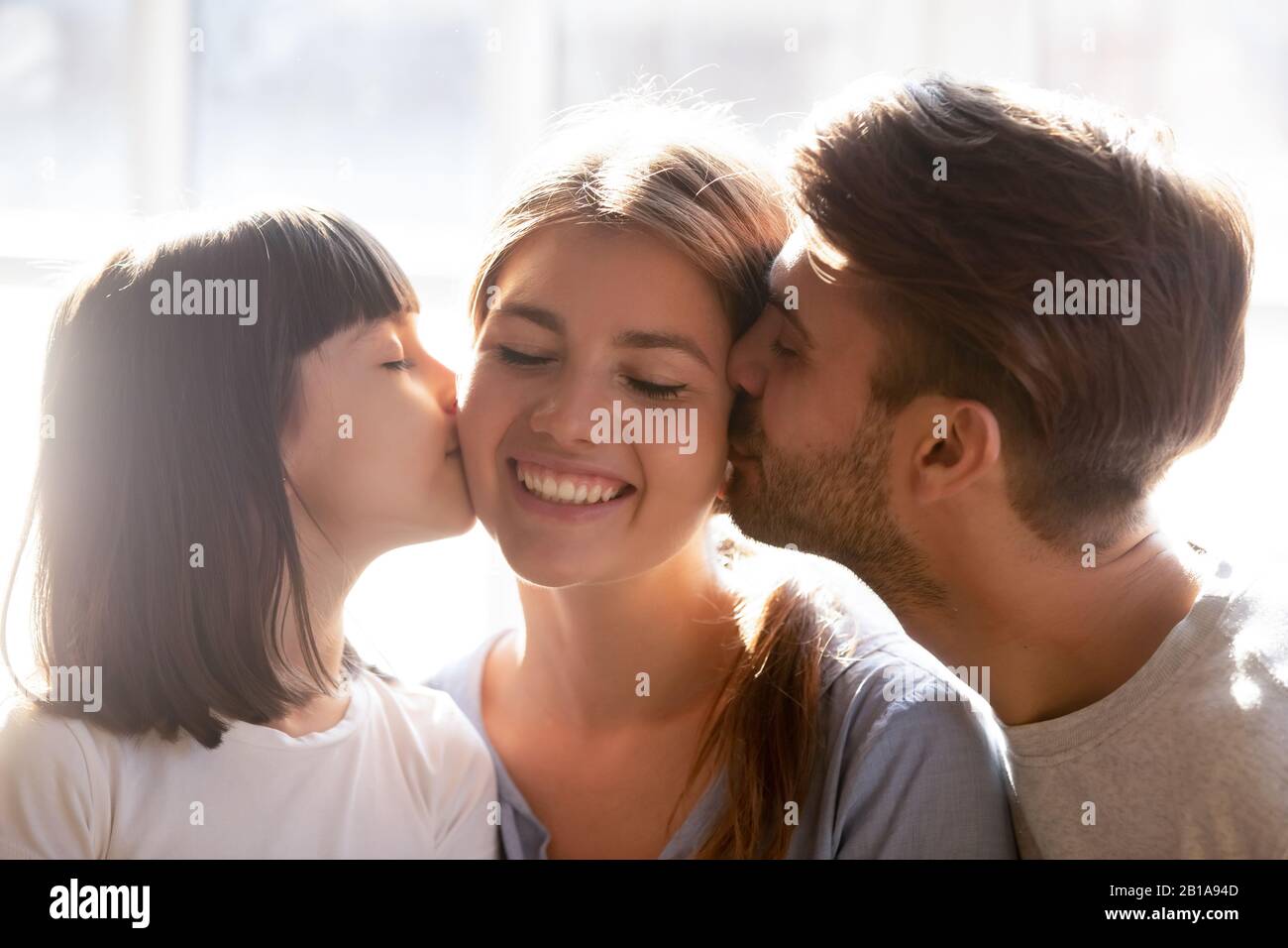 Amorevole papà e piccola figlia baciano mamma sorridente sulle guance Foto Stock