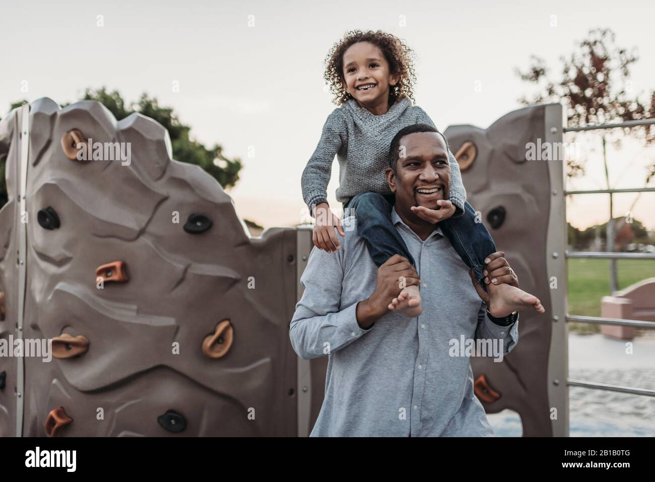 Un figlio gioioso seduto sulle spalle di un padre felice al parco giochi Foto Stock