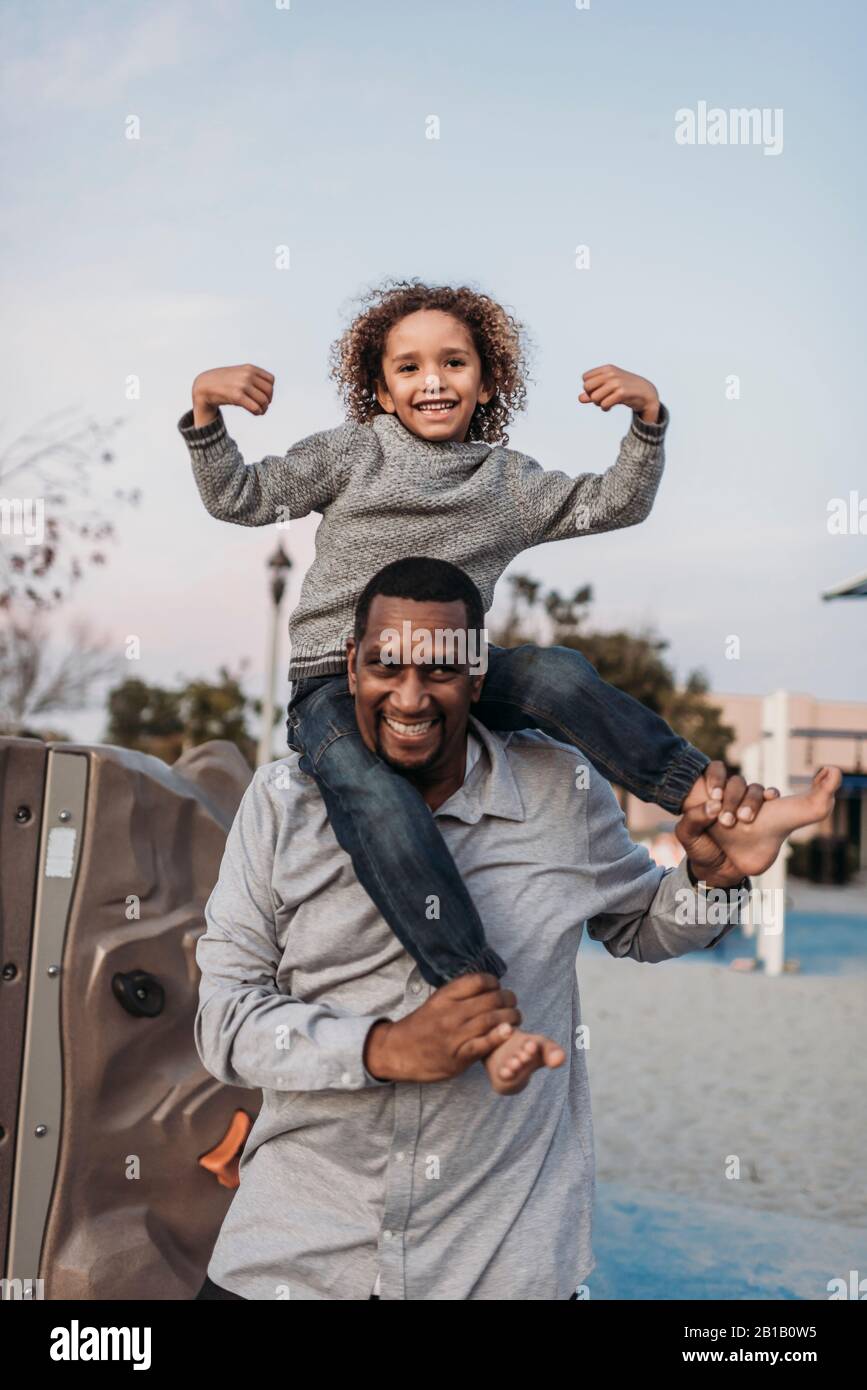 Un figlio gioioso seduto sulle spalle di un padre felice al parco giochi Foto Stock