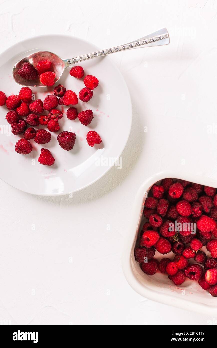Piatto bianco con cucchiaio d'argento e contenitore con lamponi. Bacche fresche estive su sfondo bianco Foto Stock