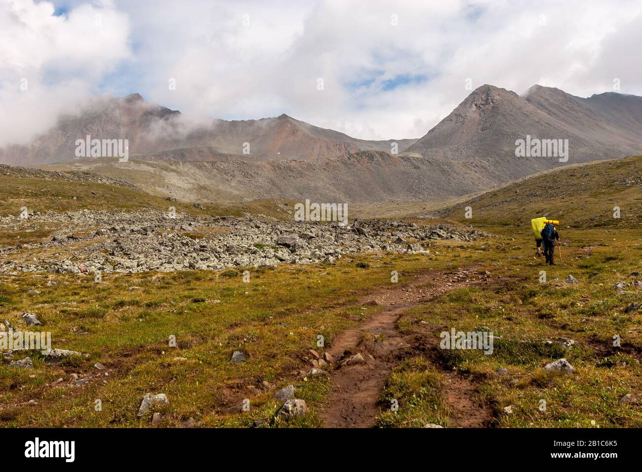I turisti con grandi zaini vanno su un sentiero escursionistico in montagna. Nuvole sopra le cime delle alte montagne. Pietra talus. Erba verde sul pendio. Foto Stock