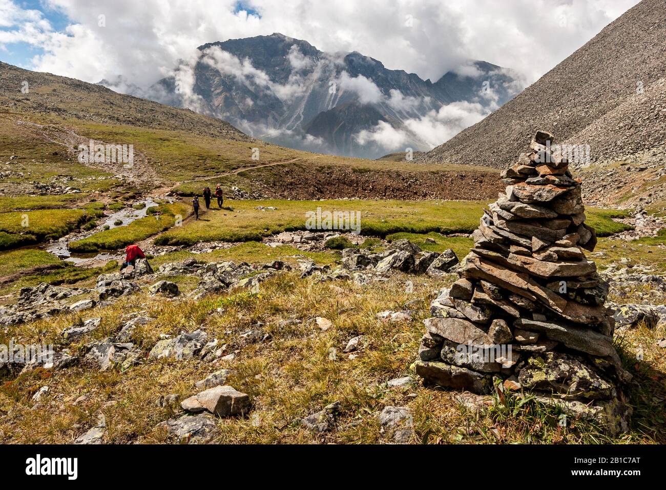 Un grande cairn in primo piano di un paesaggio montano. Nuvole bianche basse sulle montagne. I turisti con zaini percorrono il sentiero Foto Stock