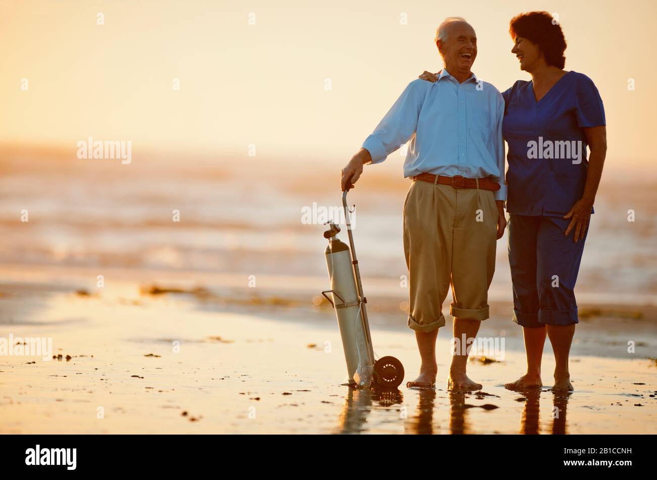 Infermiera femmina confortante senior di un paziente di sesso maschile in piedi con il suo serbatoio di ossigeno su una spiaggia al tramonto. Foto Stock