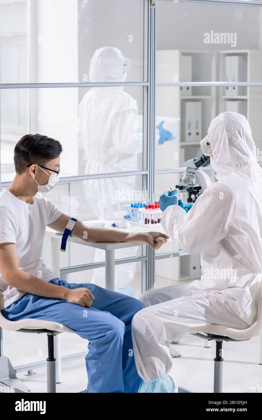 Medico in tuta protettiva che prende sangue di pazienti asiatici di coronavirus durante la ricerca medica in laboratorio Foto Stock